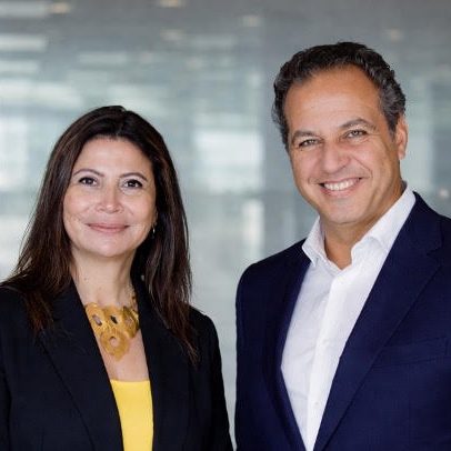 Two business colleagues, a woman in a black blazer and yellow top and a man in a navy suit, smile at the camera in a modern office setting.
