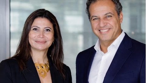 Two business colleagues, a woman in a black blazer and yellow top and a man in a navy suit, smile at the camera in a modern office setting.