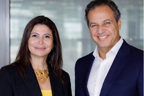 Two business colleagues, a woman in a black blazer and yellow top and a man in a navy suit, smile at the camera in a modern office setting.