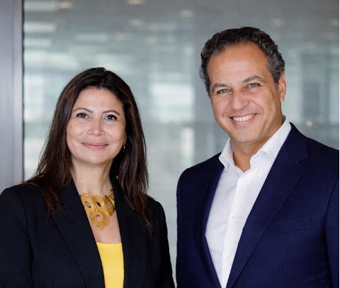 aspire-hr - Executive Search, Recruitment and Talent Management - Aspire HR Consultants Two business colleagues, a woman in a black blazer and yellow top and a man in a navy suit, smile at the camera in a modern office setting.