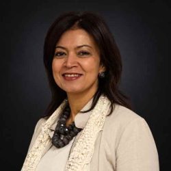 Professional headshot of a smiling woman in a beige blazer and lace scarf against a dark background, looking approachable for a corporate profile.