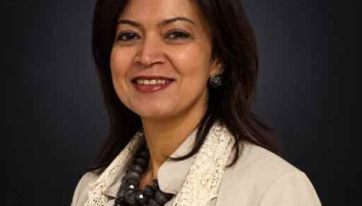 Professional headshot of a smiling woman in a beige blazer and lace scarf against a dark background, looking approachable for a corporate profile.