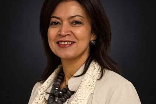 Professional headshot of a smiling woman in a beige blazer and lace scarf against a dark background, looking approachable for a corporate profile.