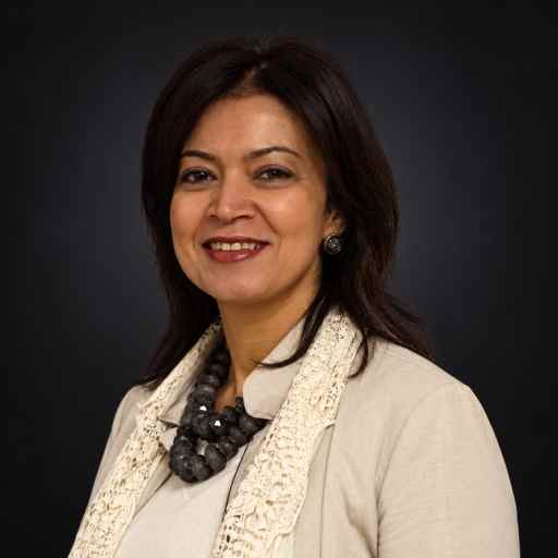 Professional headshot of a smiling woman in a beige blazer and lace scarf against a dark background, looking approachable for a corporate profile.